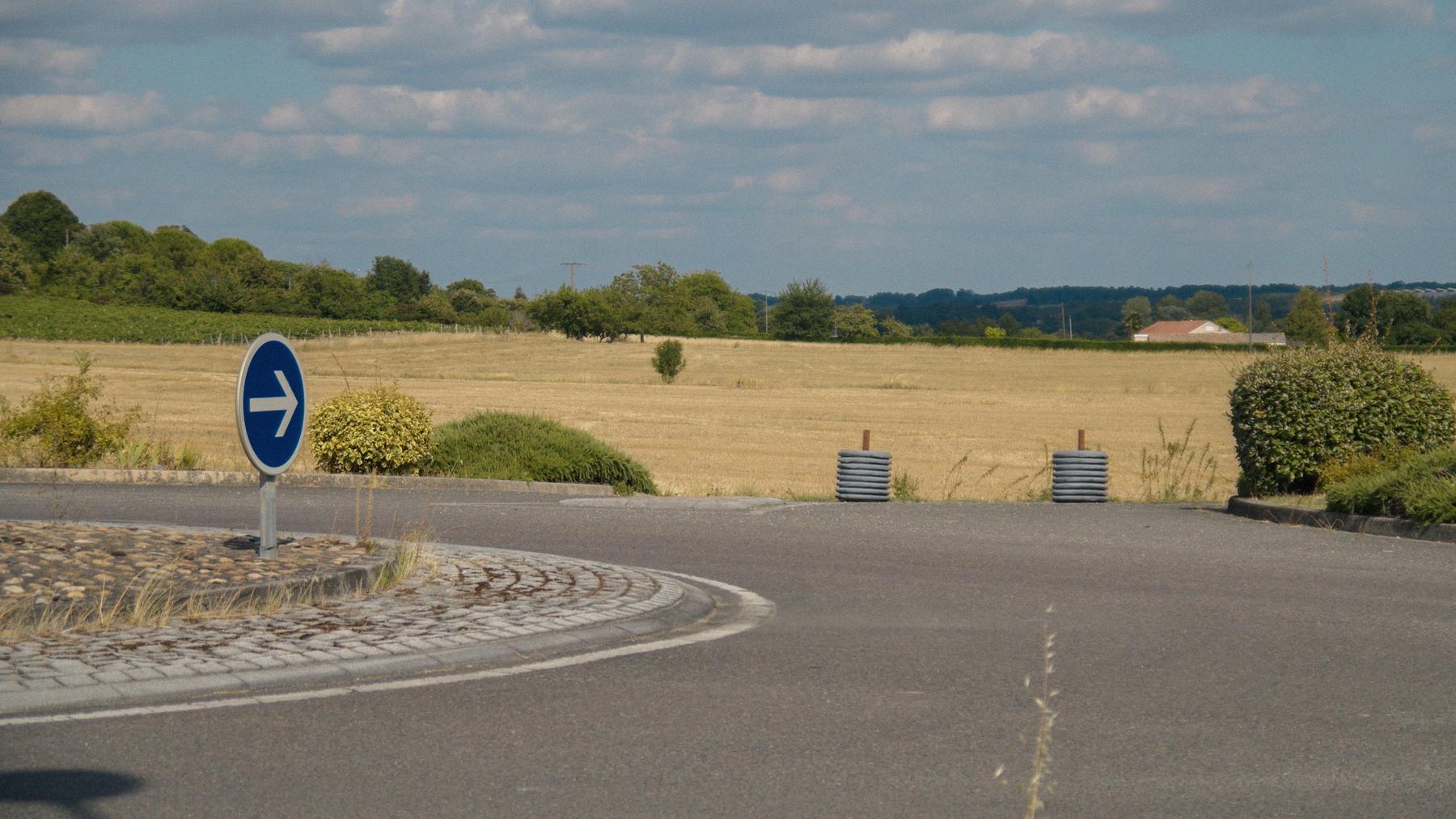 Un rond-point désert au milieu des champs moissonnés, une flèche bleue plantée au centre.