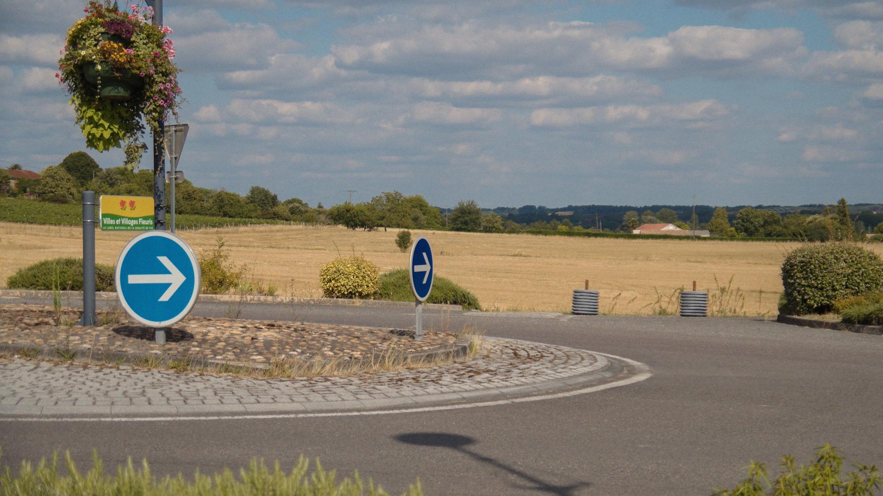 Le même rond-point fleuri, deux flèches bleues sous un ciel de petits nuages.