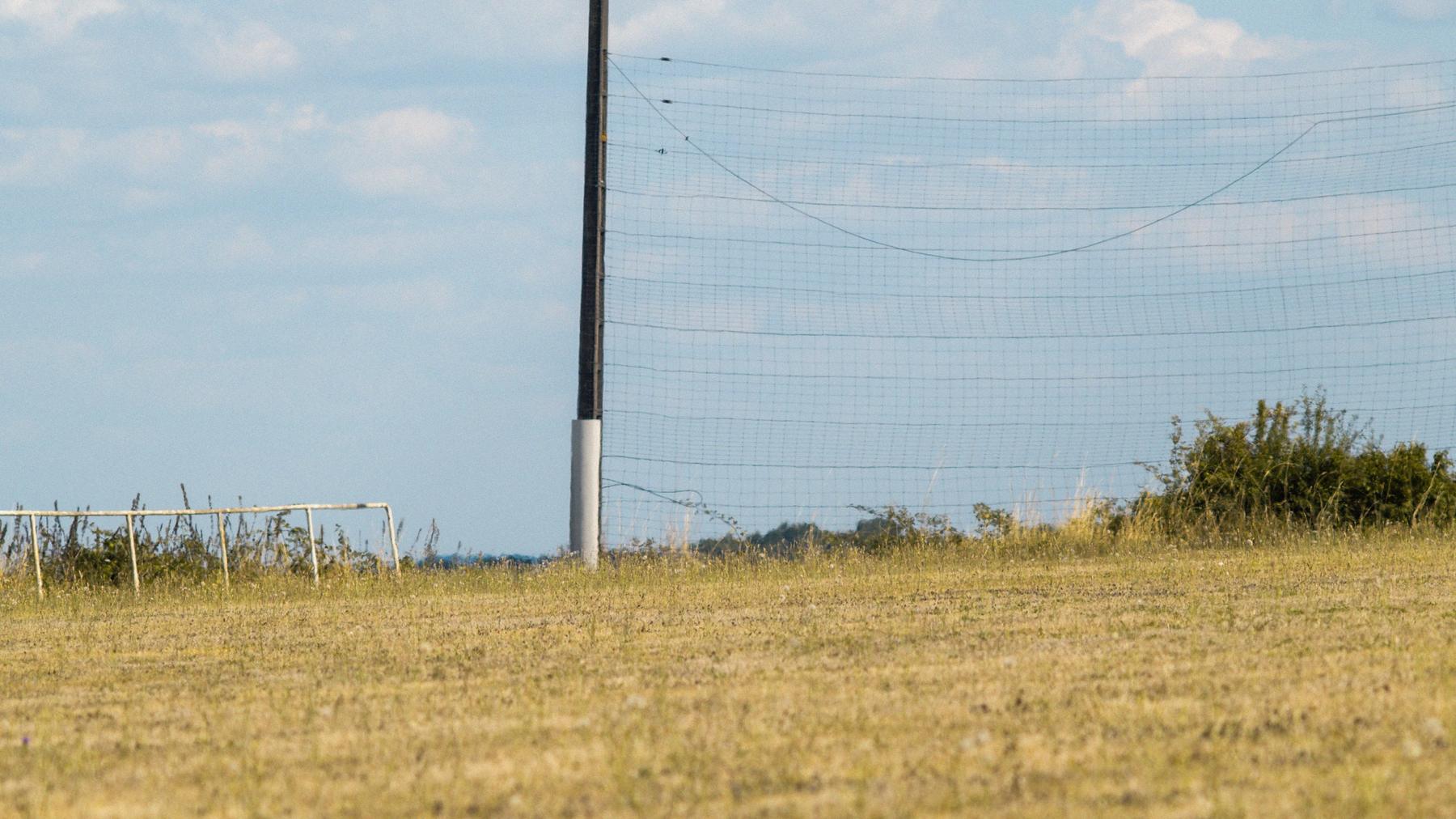 Un poteau et un filet pare-ballons au bord d'un terrain de football grillé par l'été.